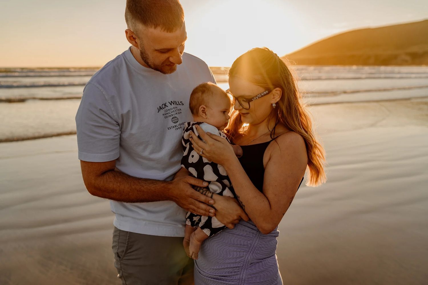Family embraces on a sandy beach against a dramatic sunset backdrop, with waves gently rolling in.