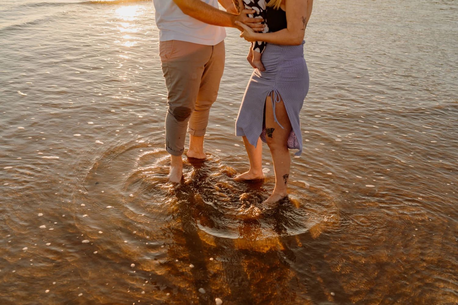 People standing in shallow ocean water during golden hour create intimate silhouettes against the setting sun.