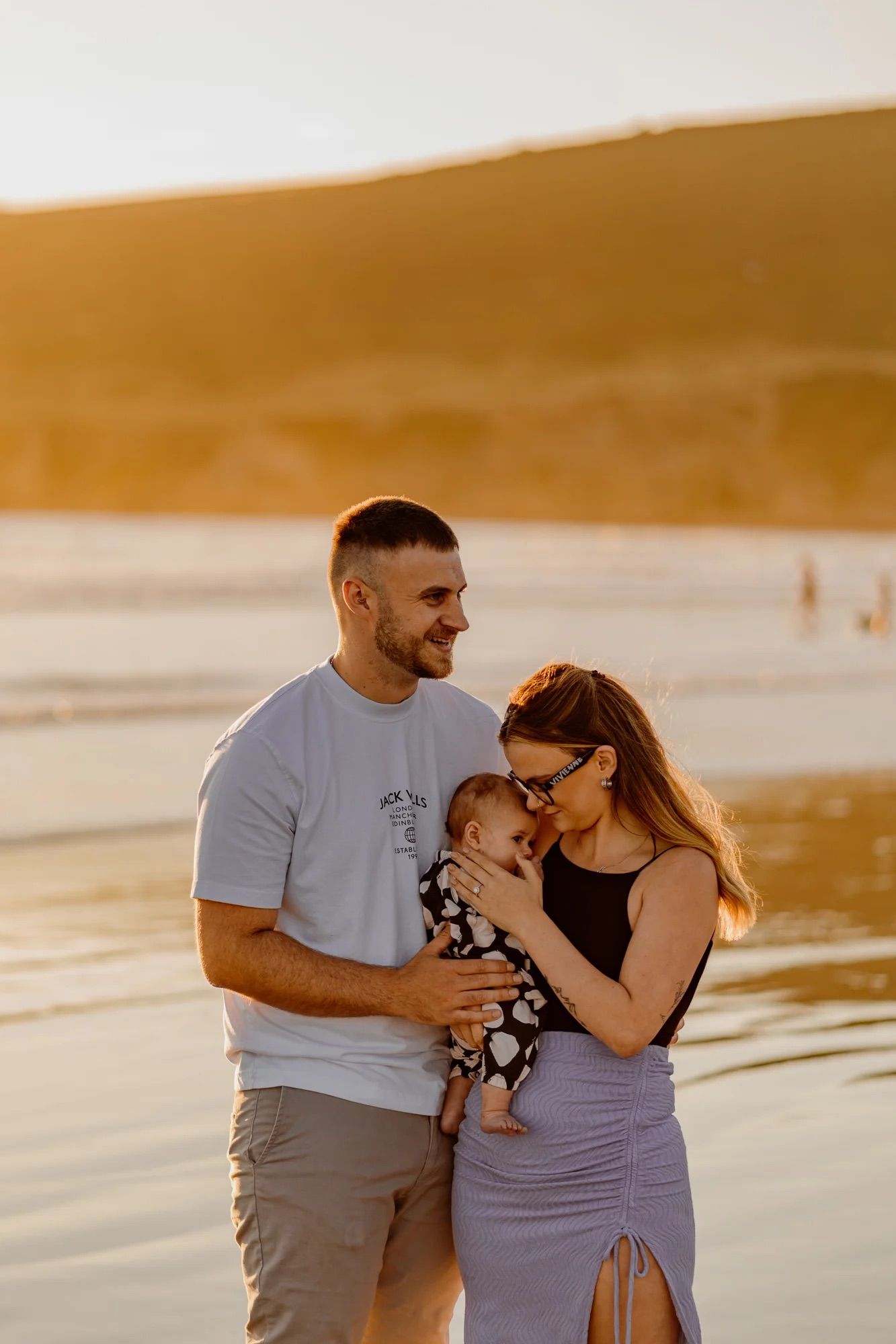 A couple shares tender moments on a beach at sunset while wearing casual clothes in warm golden light.