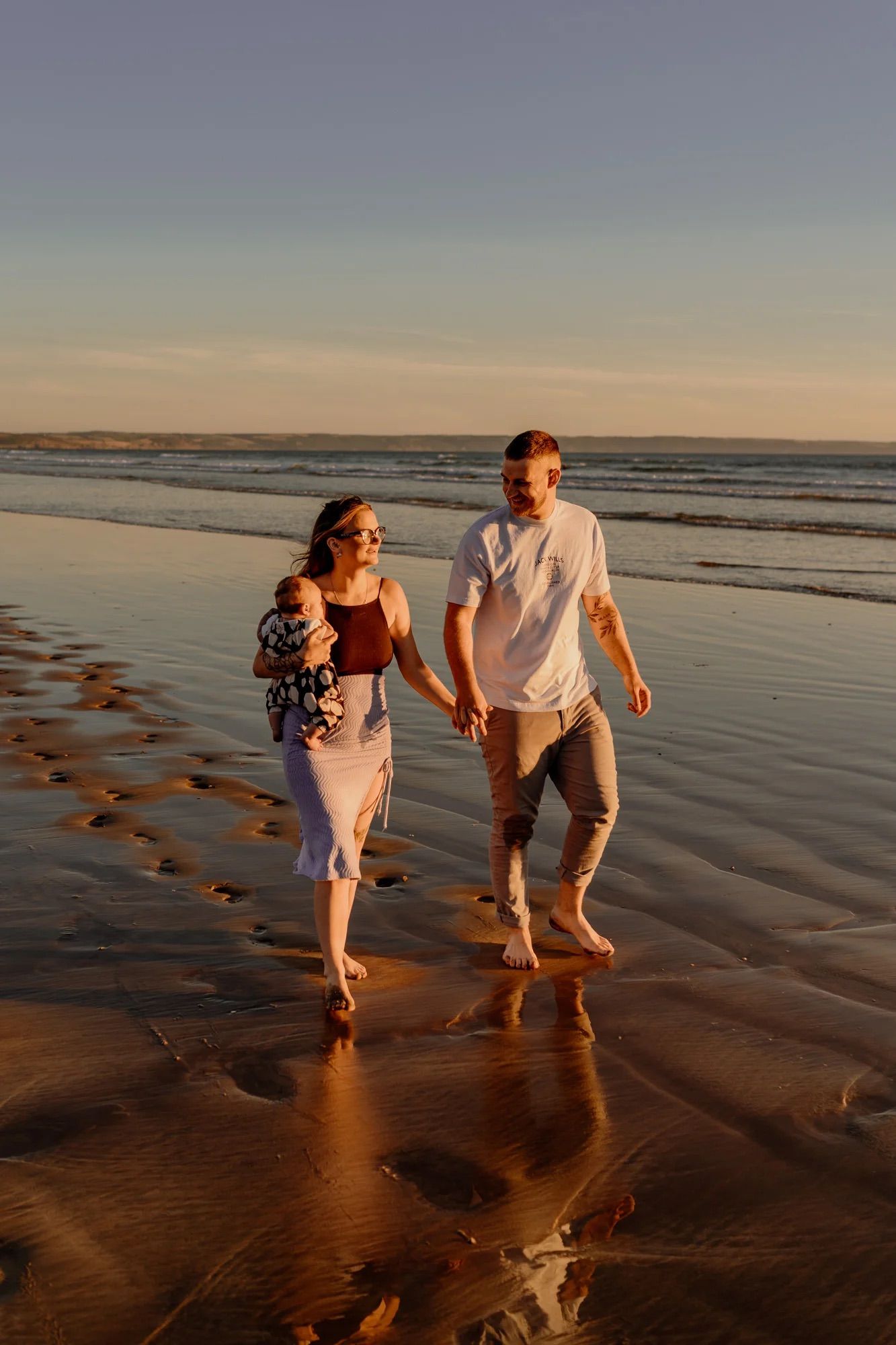 Two people strolling together on a wet sandy beach during a beautiful orange sunset with footprints trailing behind.