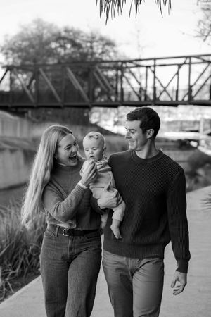 Black and white photo series of a couple holding their baby while walking along a path near a bridge.