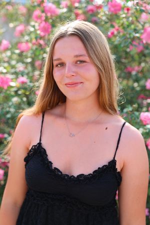 Someone wearing a black lace-trimmed dress poses in front of blooming pink roses in a garden setting.