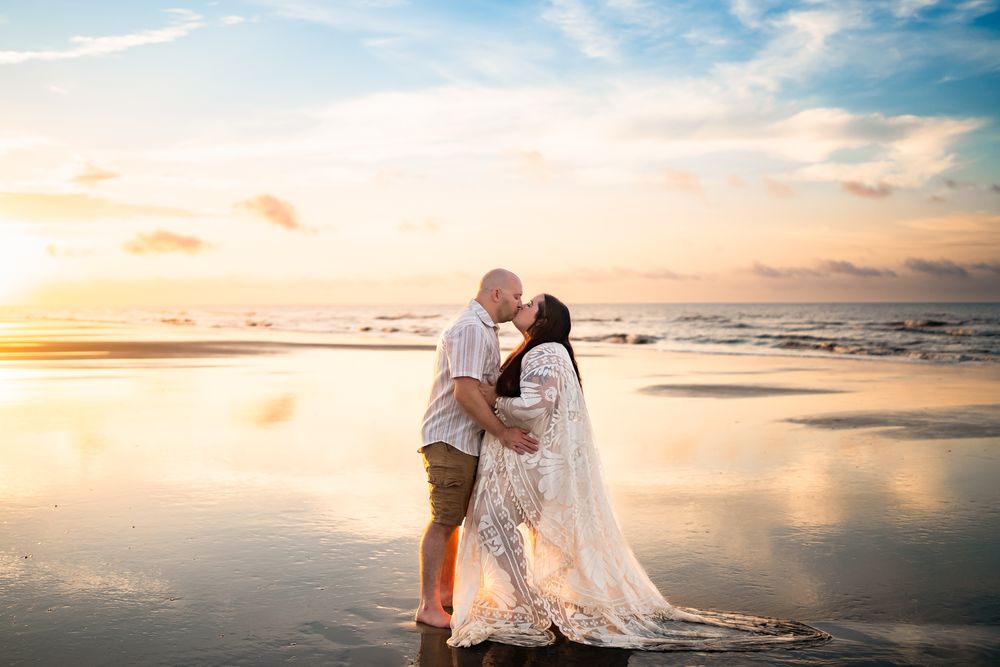 Couple in wedding attire embracing on a scenic beach at sunset with ocean waves and pastel sky in the background.