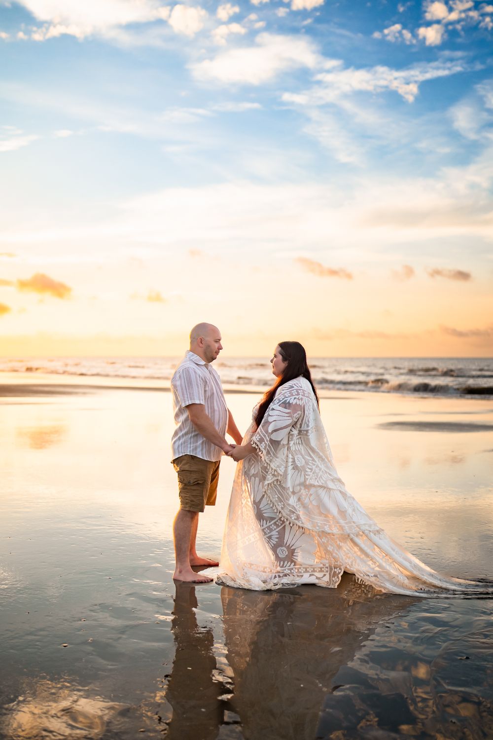 Two people share a romantic moment at sunset on a reflective beach with dramatic clouds and golden light.