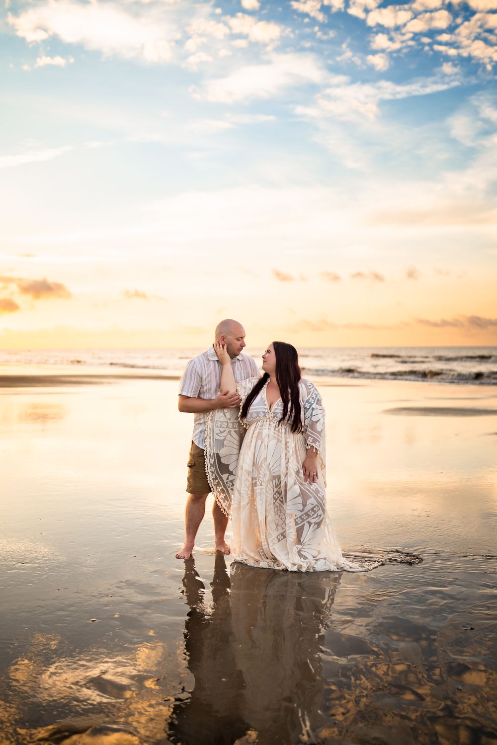 Two people embrace on wet sand at the beach during a beautiful golden sunset.