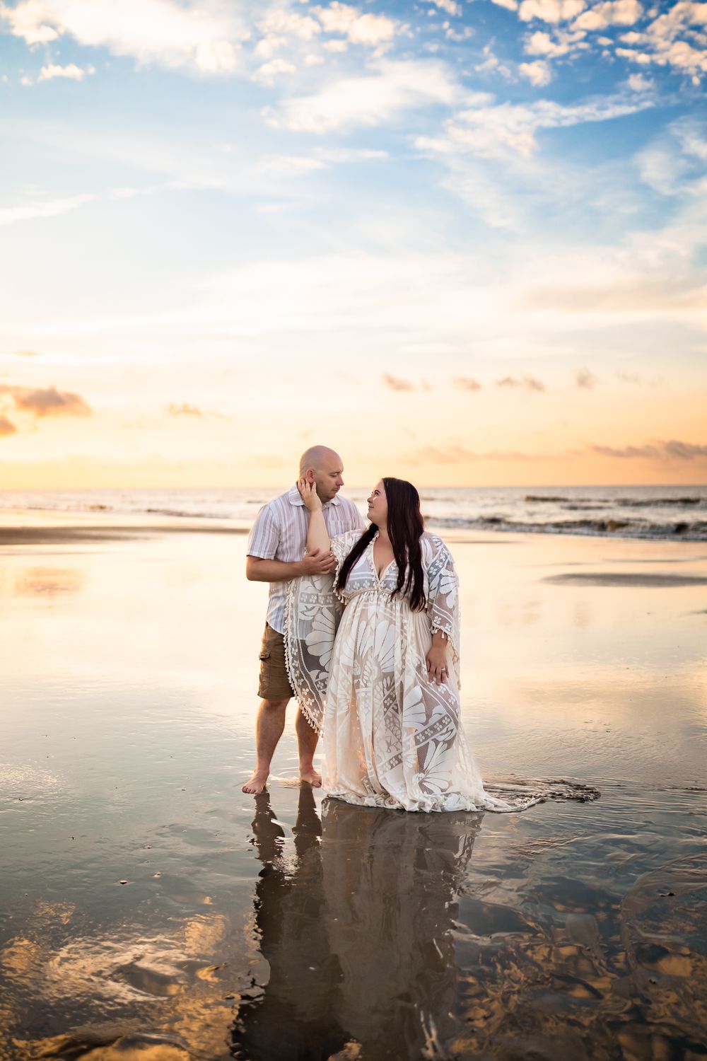 A romantic couple shares an intimate moment on a reflective beach at sunset with golden light bouncing off the wet sand.