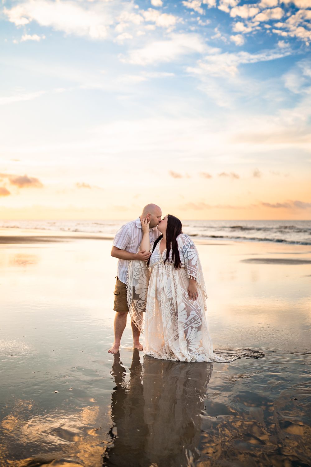 A couple shares an intimate moment on a reflective beach at sunset with soft golden light and gentle waves.