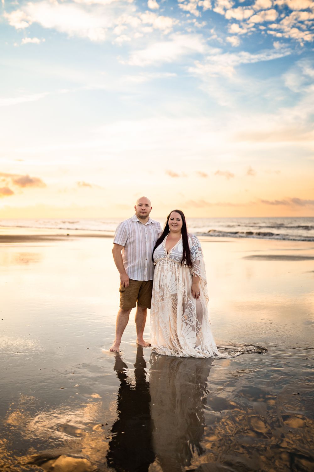 A couple stands together at sunset on a reflective beach with golden light and clouds in the sky.