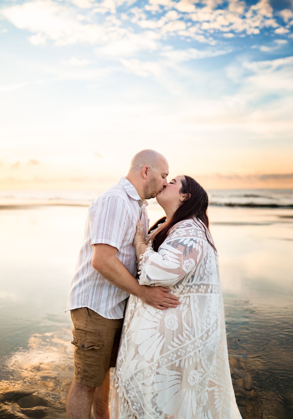 A romantic beach photoshoot at sunset with the couple embracing on wet sand with golden light.