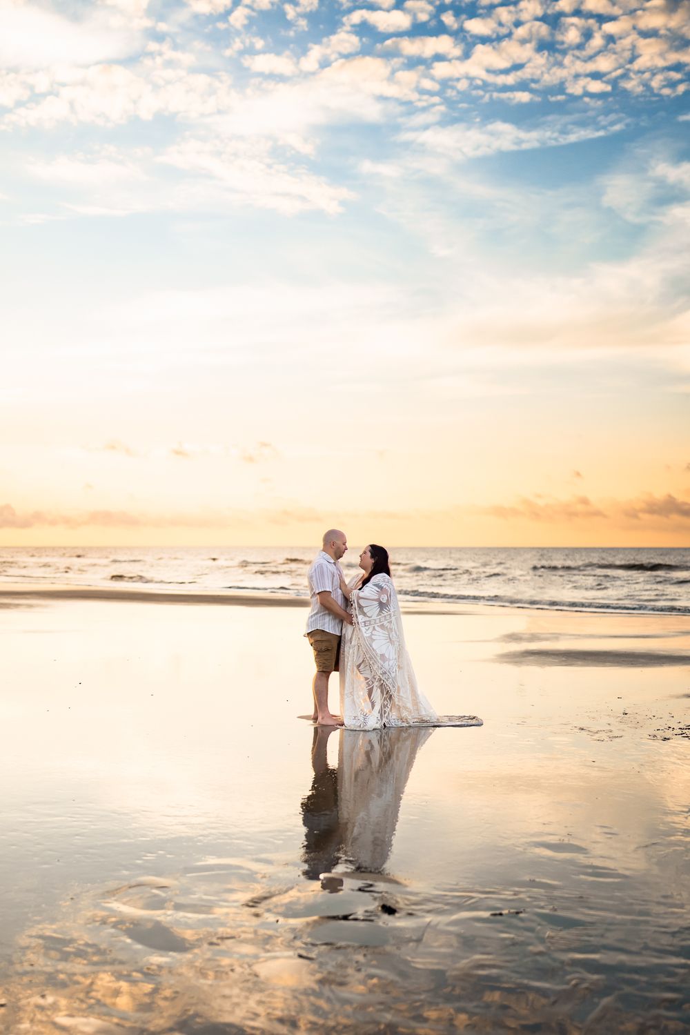 Silhouettes of a couple share an intimate moment on the beach with sunset reflections on the wet sand.
