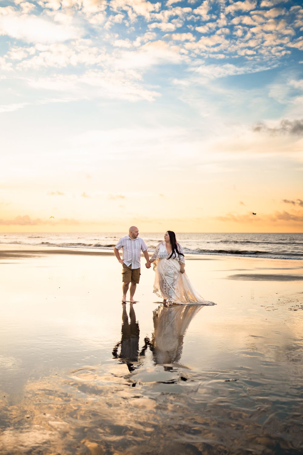 Couple walking on wet sandy beach at sunset with dramatic cloudy sky and ocean reflections creating romantic silhouettes.