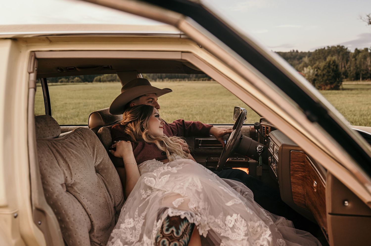 A couple shares a tender kiss inside a vintage car during their wedding celebration in a scenic countryside setting.