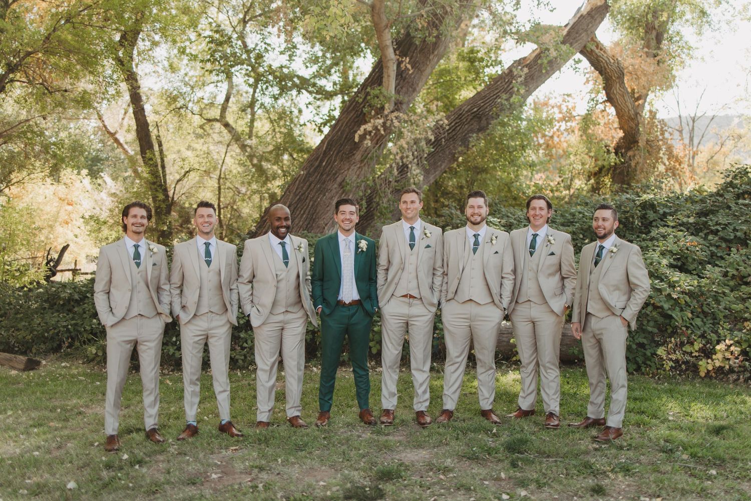 Wedding party group stands in alternating formation outdoors wearing light gray suits beside evergreen trees.