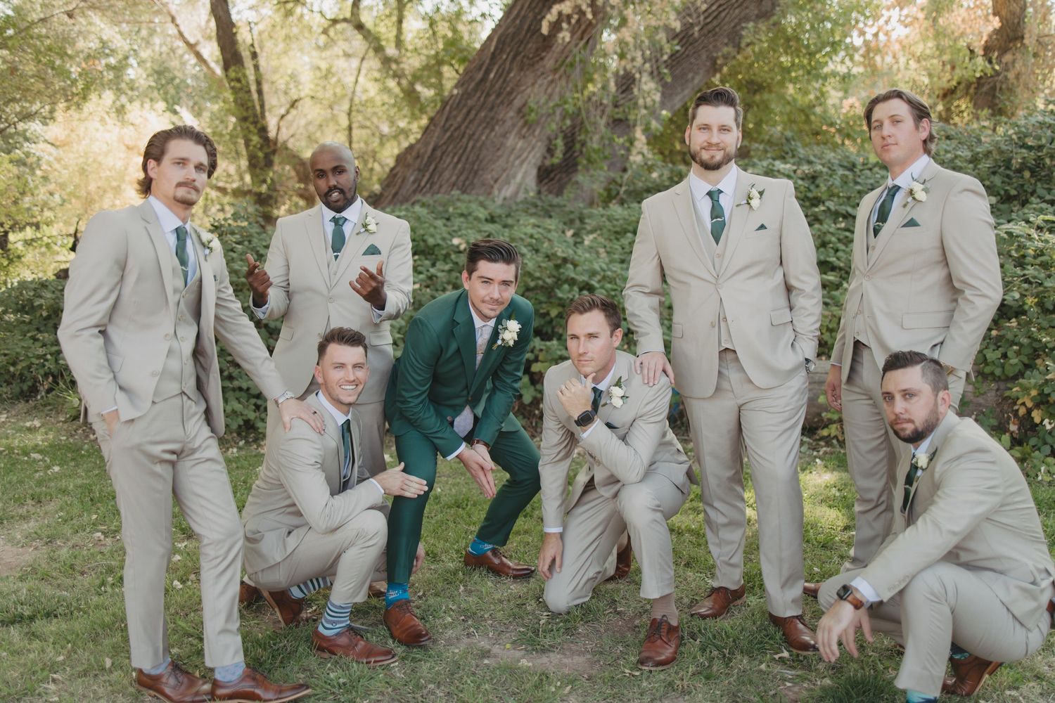 Groomsmen in light beige suits and blue ties pose casually on grass during outdoor wedding photoshoot.