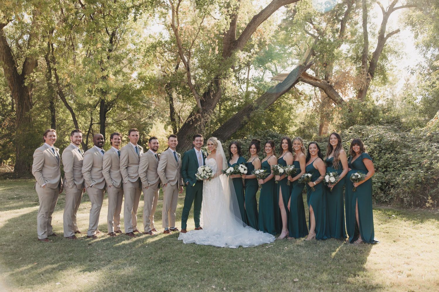 Wedding party members in green and gray attire pose together outdoors for formal group photos in a natural setting.