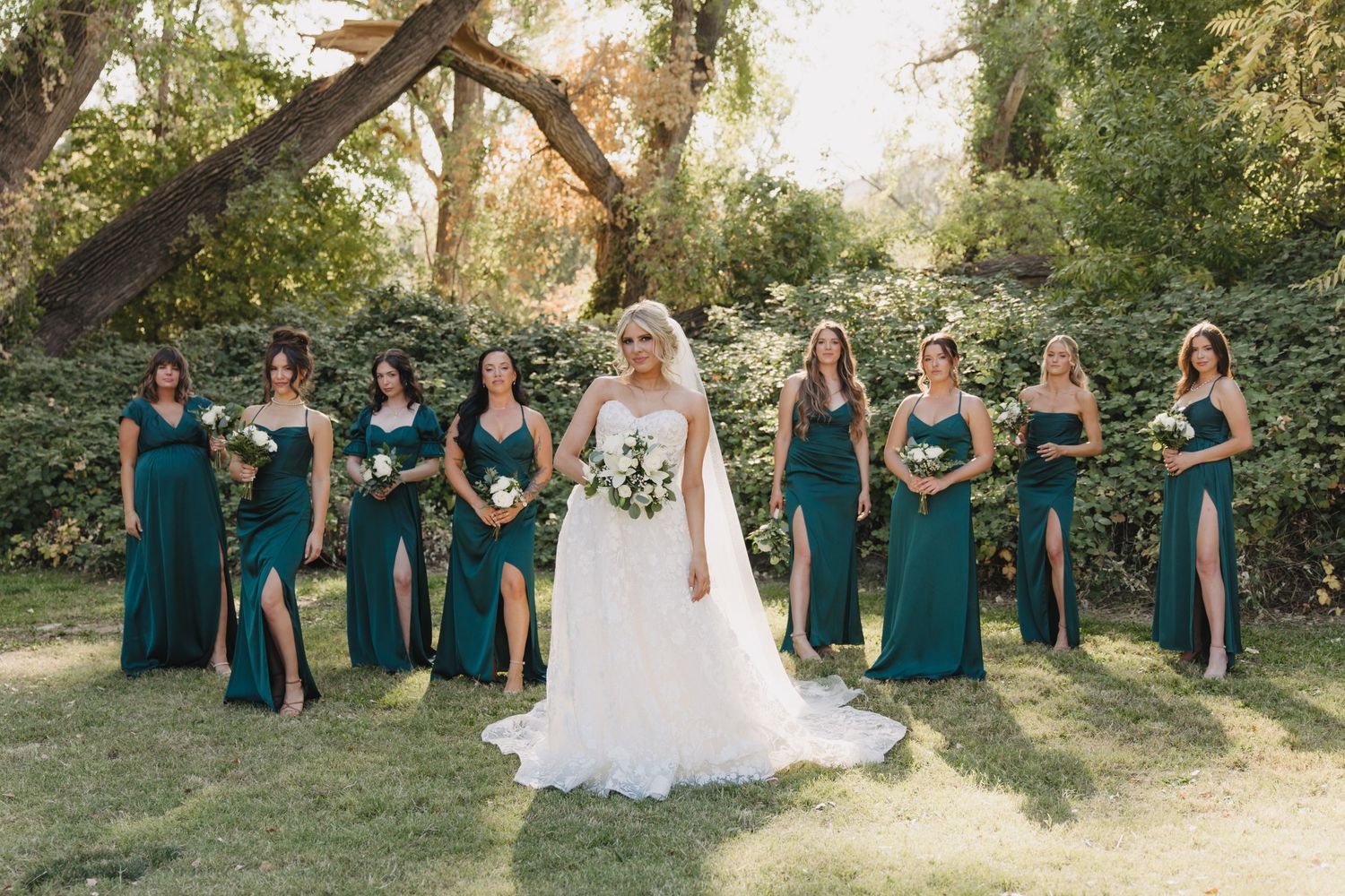 Wedding party wearing elegant emerald green bridesmaid dresses and white bridal gown pose together in outdoor garden setting.