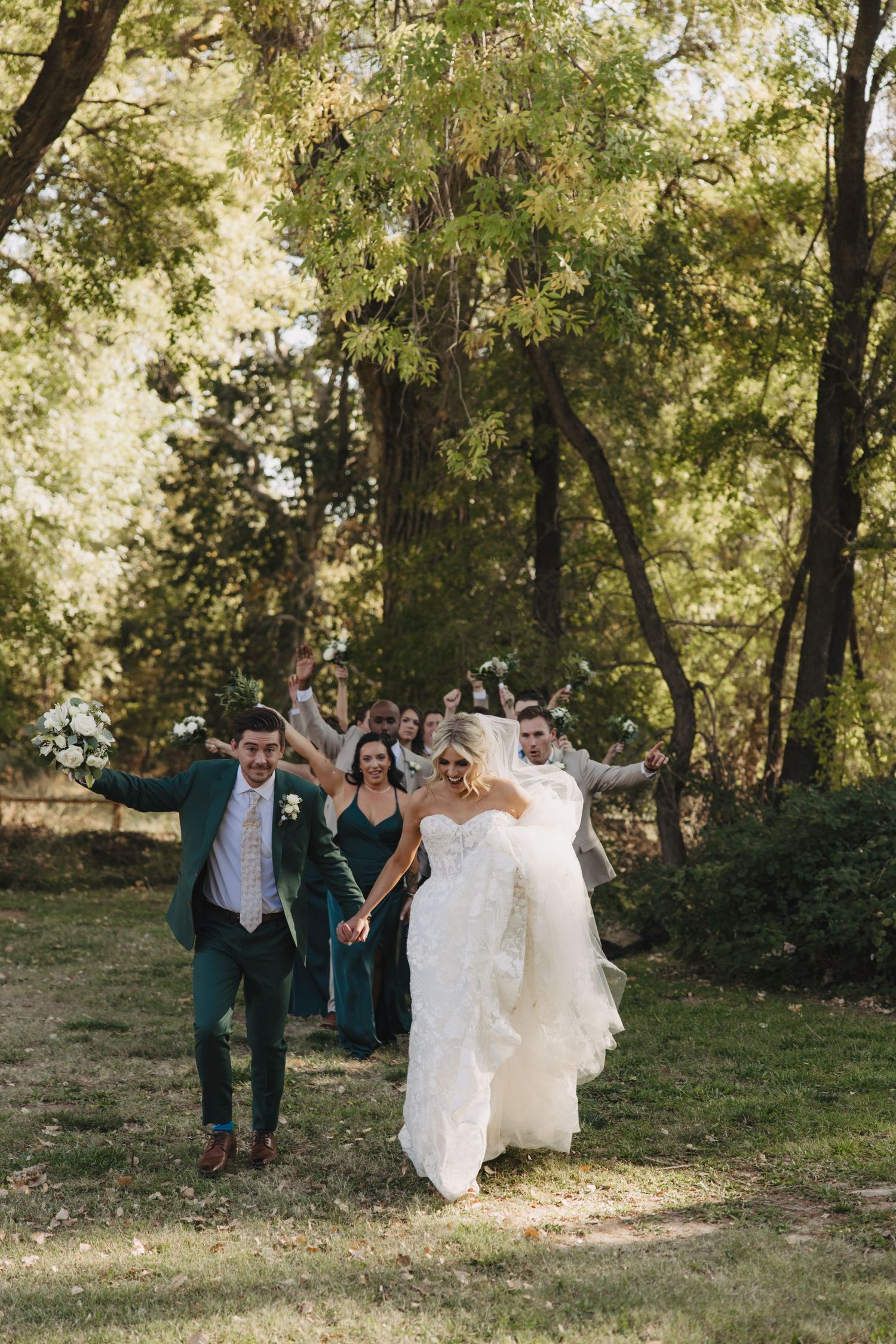 Wedding party members dash joyfully through a scenic wooded area on a bright sunny day.