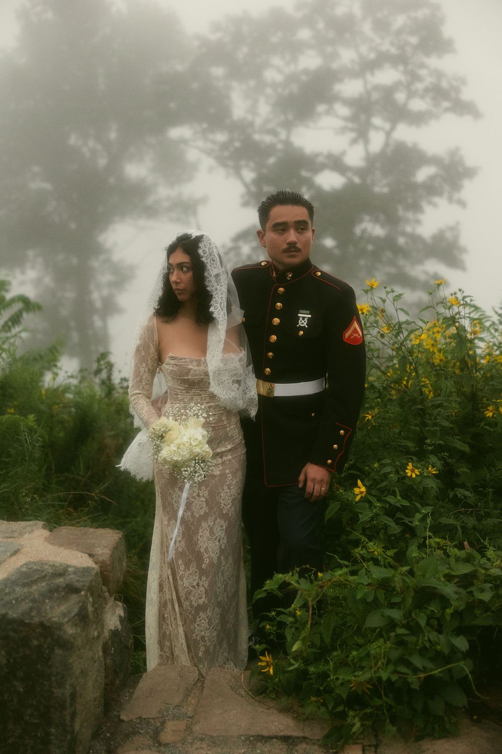 Couple in wedding attire posing together on a misty path with yellow flowers in the background.