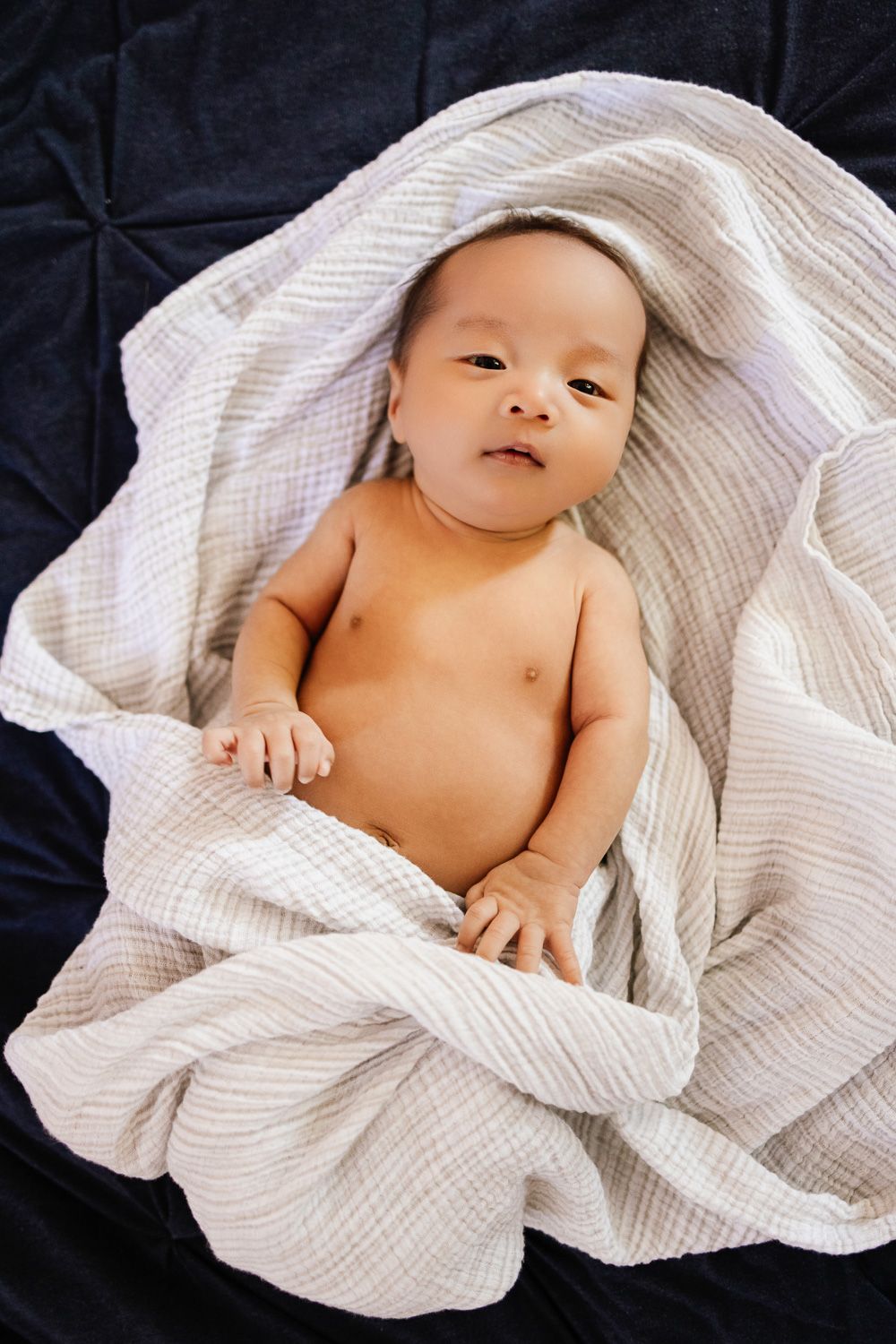 A newborn baby boy smiles at the camera while partially wrapped in a swaddled during his in-home newborn session by Cindy Tucey Photography