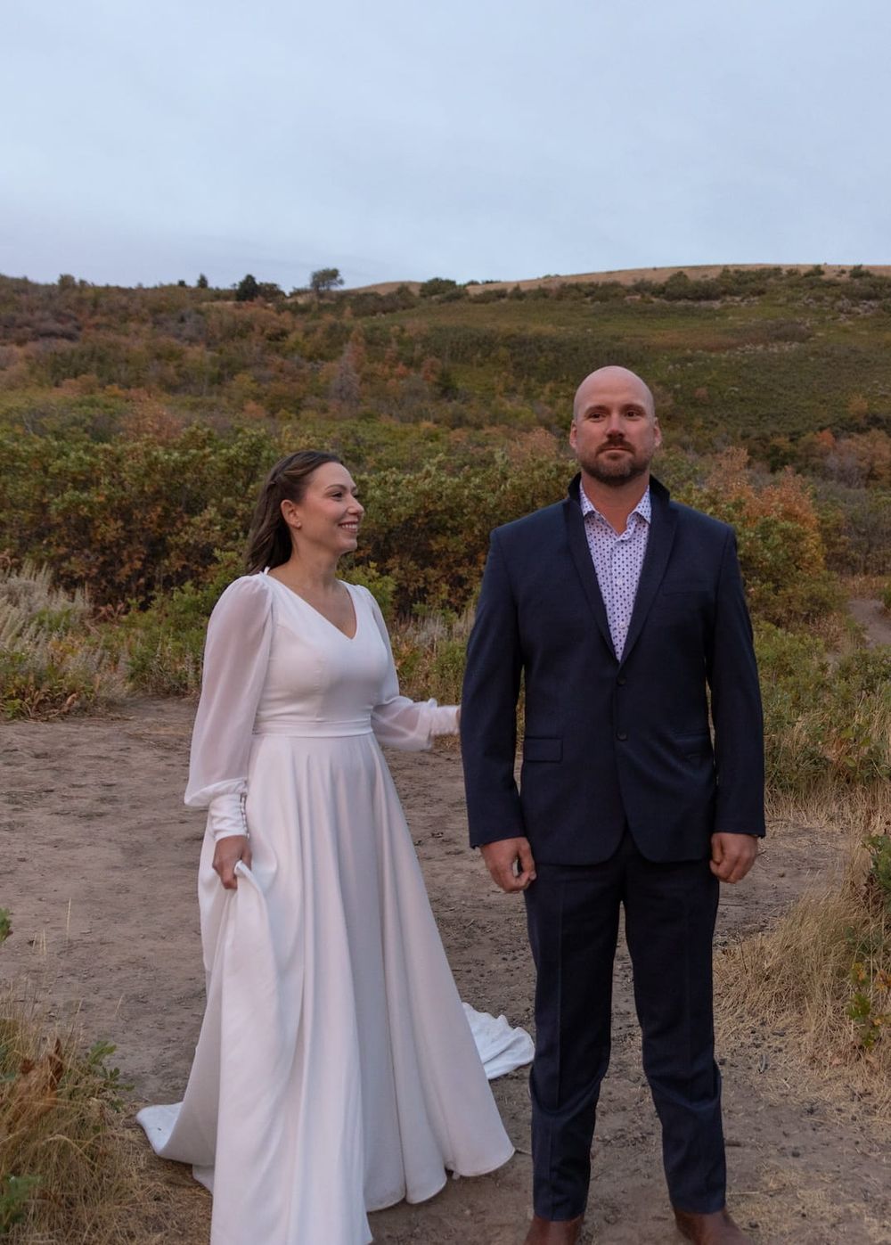 A couple in contrasting formal attire poses together on a hillside path during autumn.