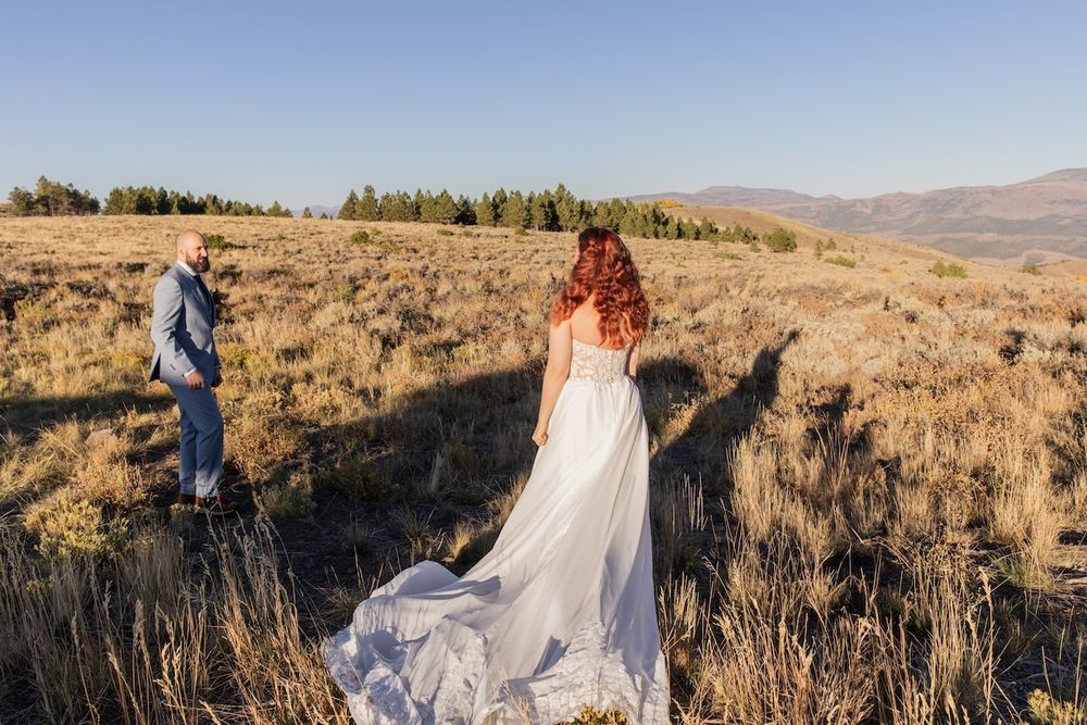 A photographer captures a shot of someone in a flowing white dress with red hair standing in a golden meadow at sunset.