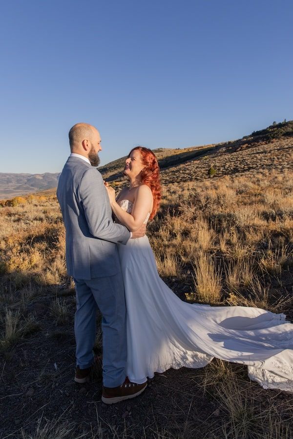 Romantic couple embraces in a scenic desert landscape during sunset with bride's flowing white dress trailing on ground.