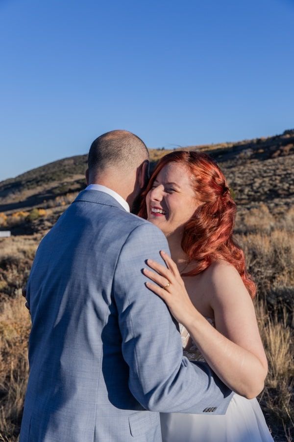 A romantic couple shares a tender embrace against a desert mountain landscape in soft sunlight.