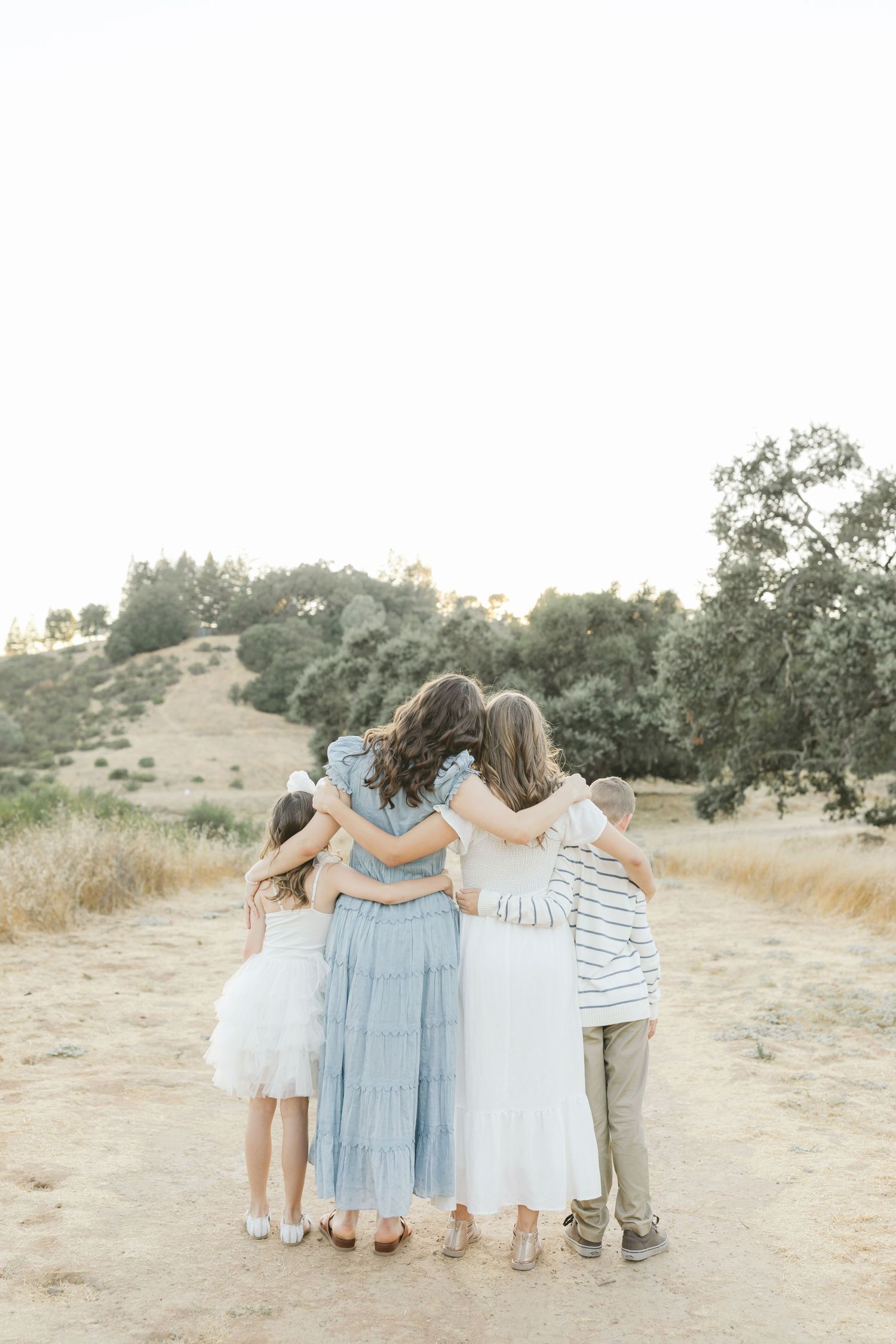 A group stands together with arms around each other on a dirt path overlooking hills at sunset wearing light colored clothing.Film by Chelse