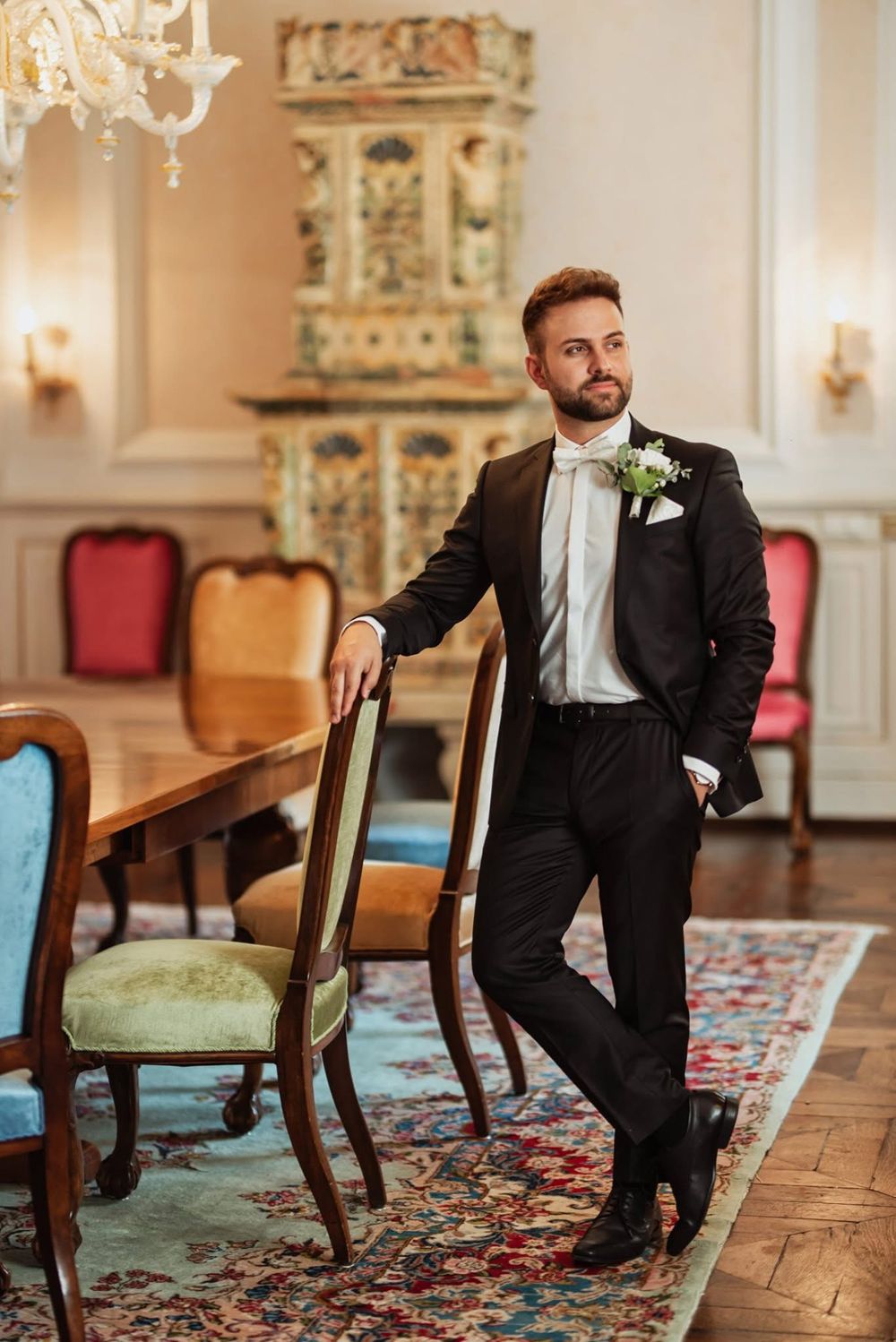 A groomsman in a black tuxedo poses playfully in a vintage ballroom with ornate decor and colorful patterned carpet.