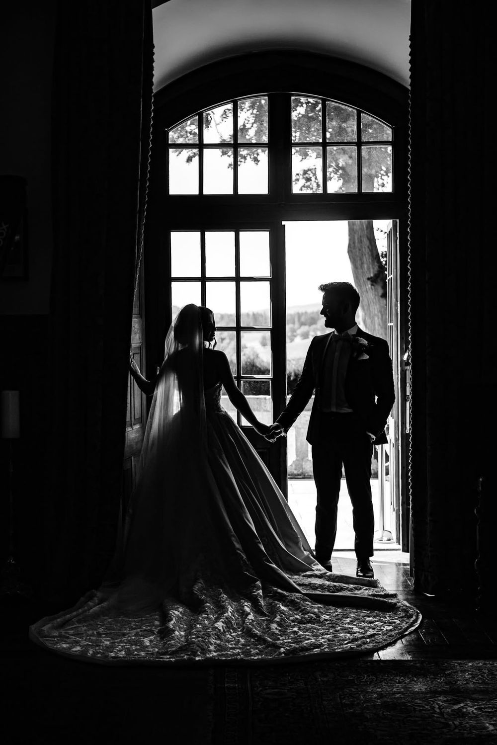 Black and white silhouette wedding photos showing couple in front of large arched window in dramatic lighting.