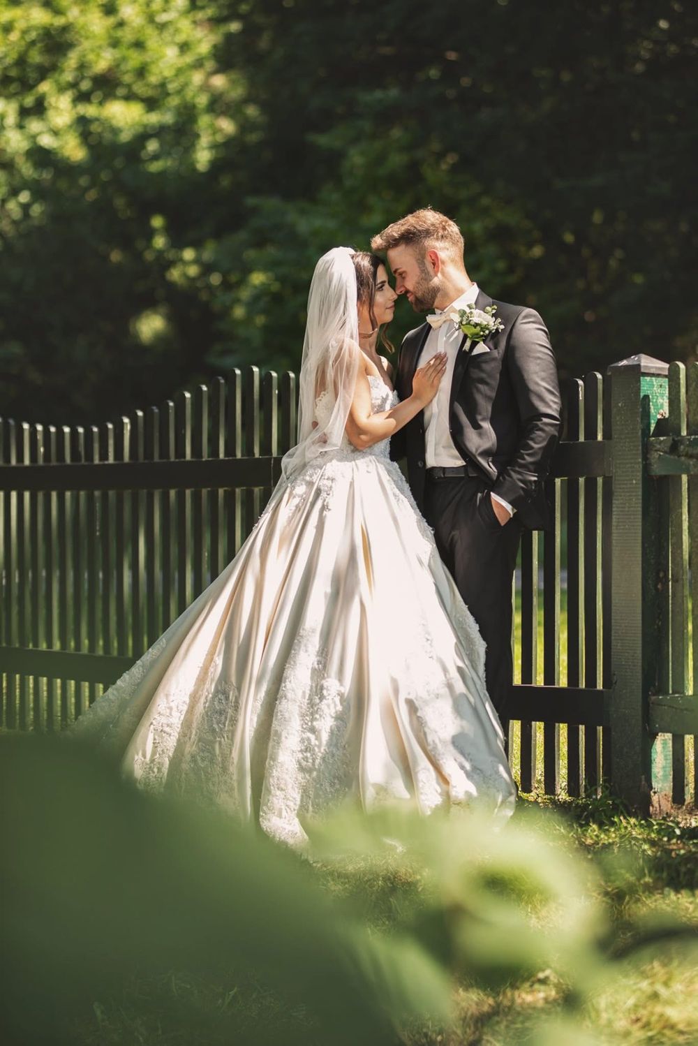 Romantic wedding couple shares tender moment together in lush garden setting with sunlit greenery and wooden fence.