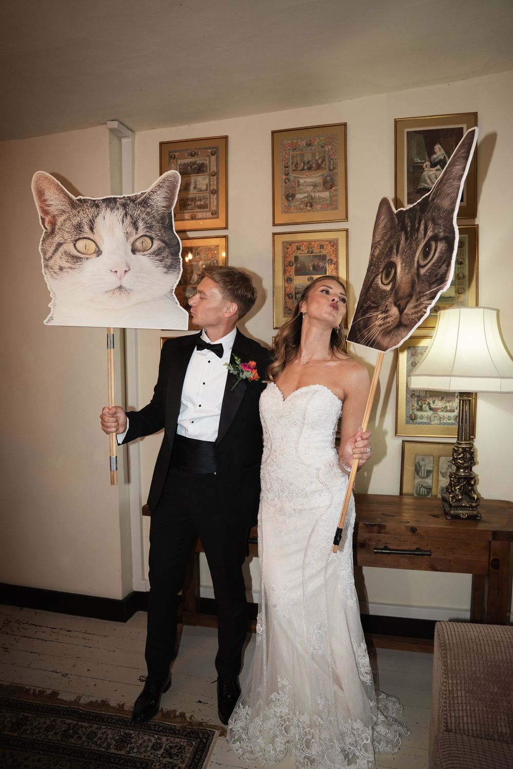 Two people in formal wedding attire share a playful moment posing together against a wall with framed artwork.