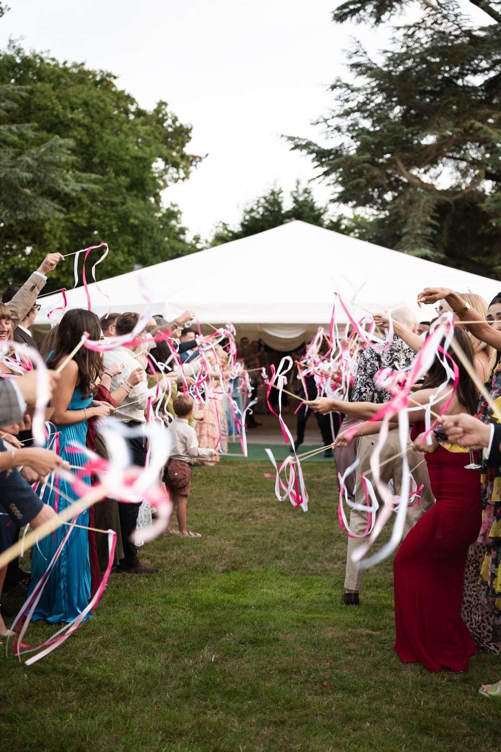 Wedding guests waving pink and white ribbon streamers in celebration on a grassy outdoor lawn.