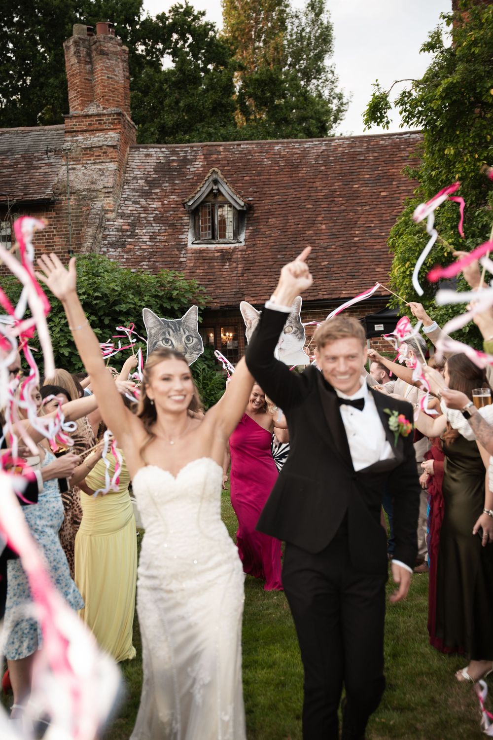 Wedding party celebrates outside historic brick building by waving pink ribbon wands in festive celebration.