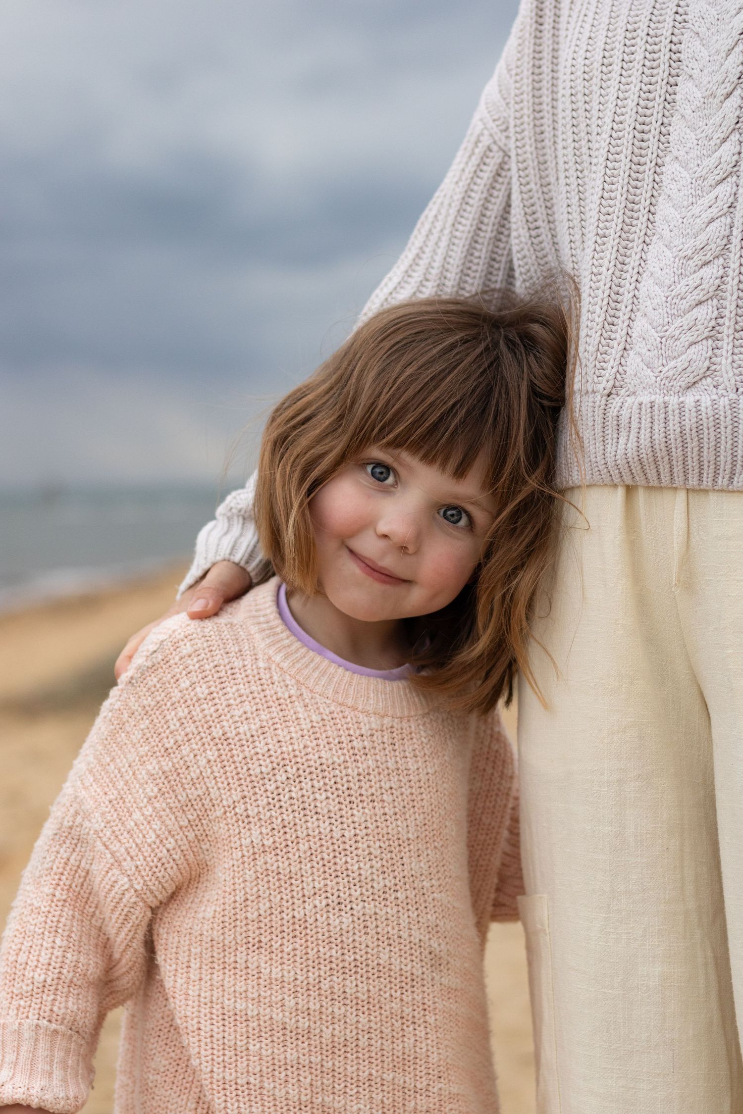 Small child wearing a pink knit sweater peeks playfully from behind an adult wearing a white knit sweater.