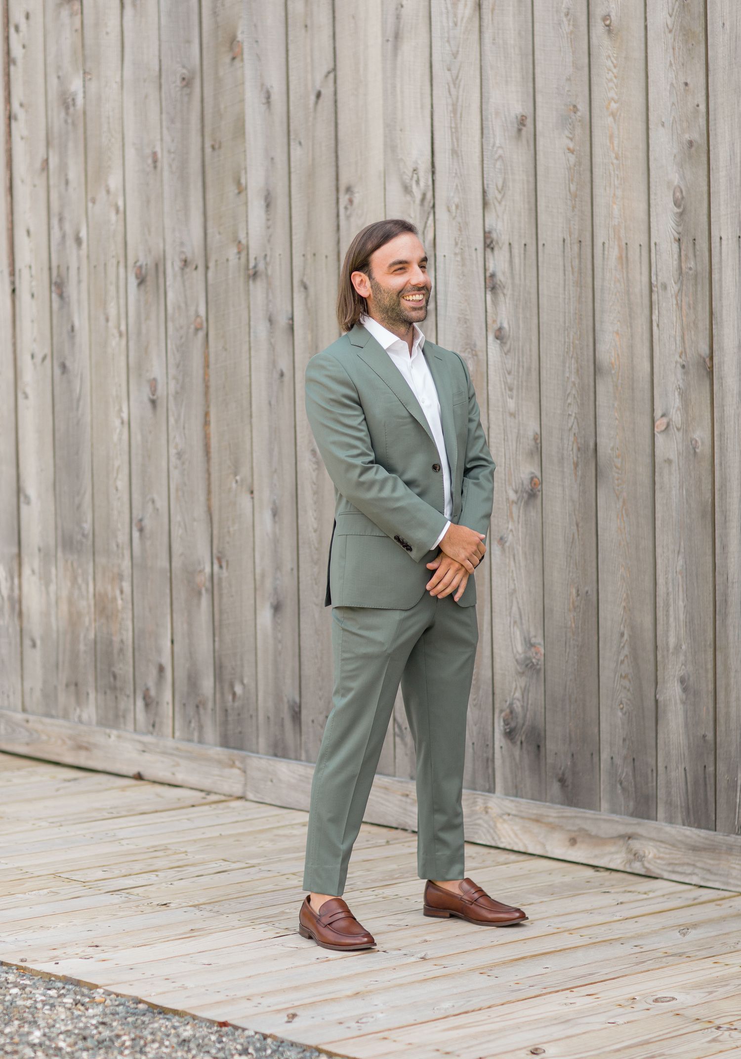 A person in a sage green suit and brown leather shoes stands on a wooden deck against a weathered wood fence.