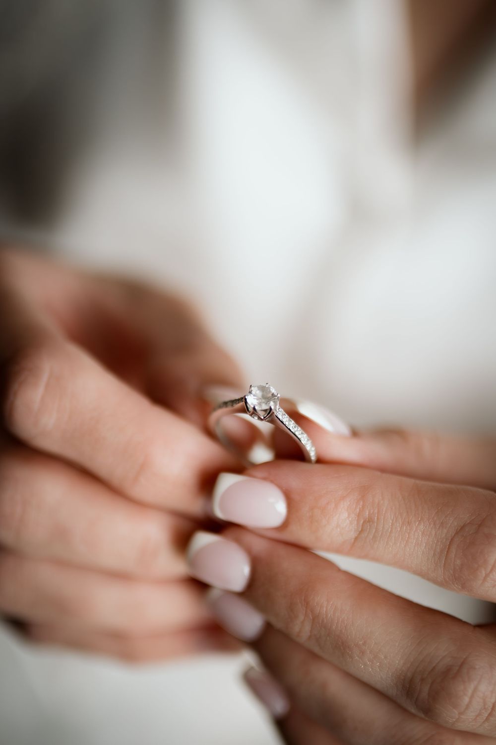 Close-up of hands holding a diamond solitaire engagement ring with pale pink manicured nails.