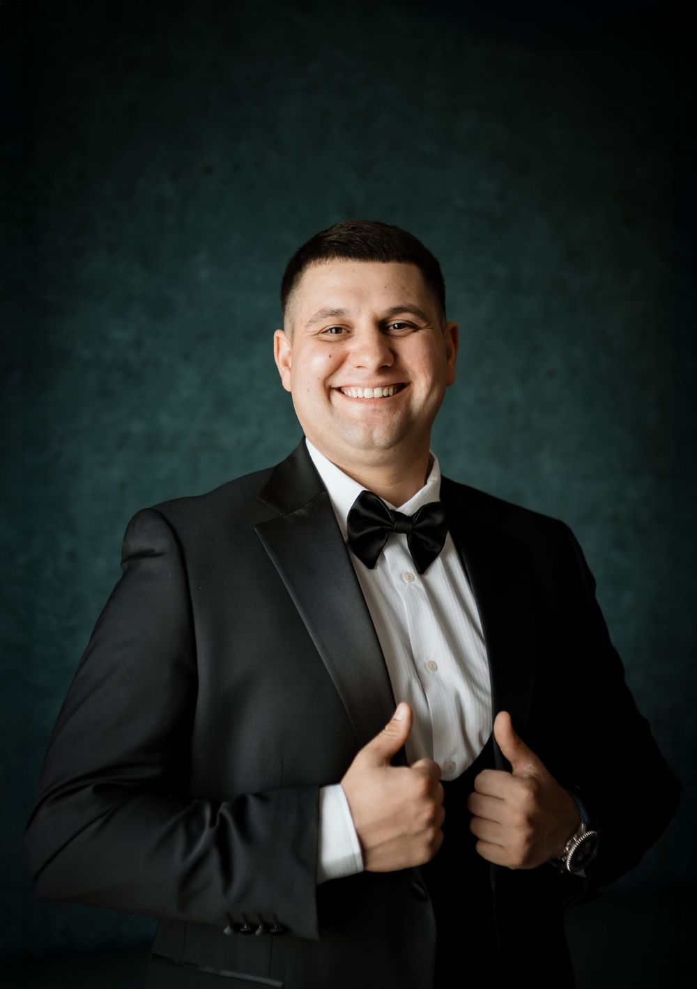 Professional portrait with black tuxedo and bowtie against a textured dark green backdrop.
