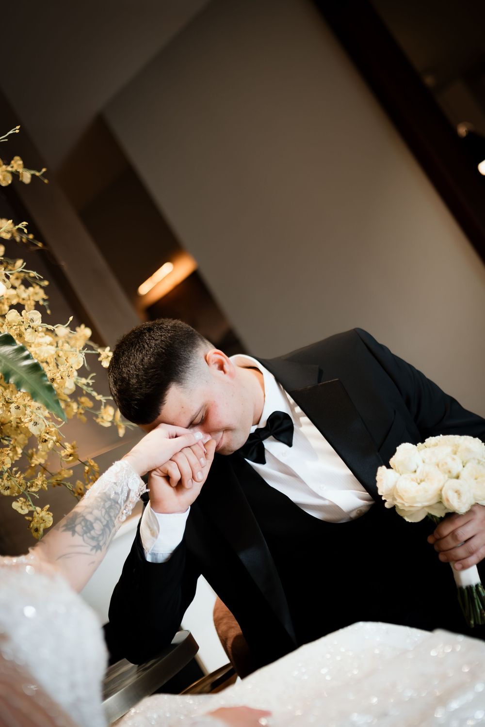 An emotional wedding moment captured as the groom kisses the bride's hand while holding a white bouquet.