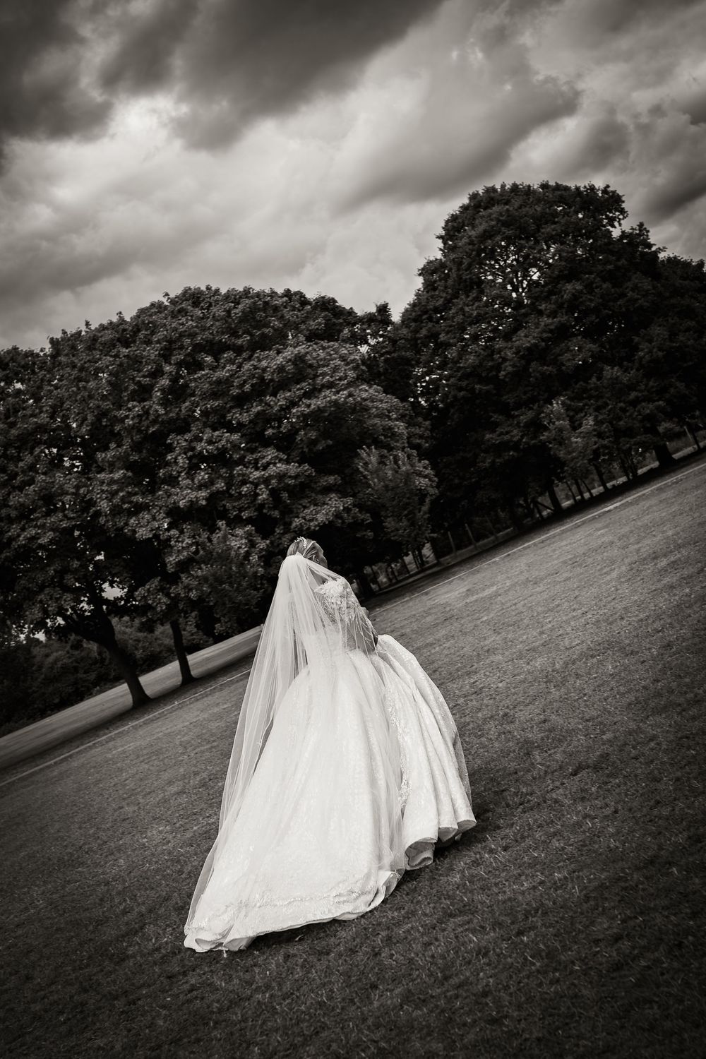 A dramatic black and white photo of a bride in flowing white dress walking on a hillside under stormy skies.