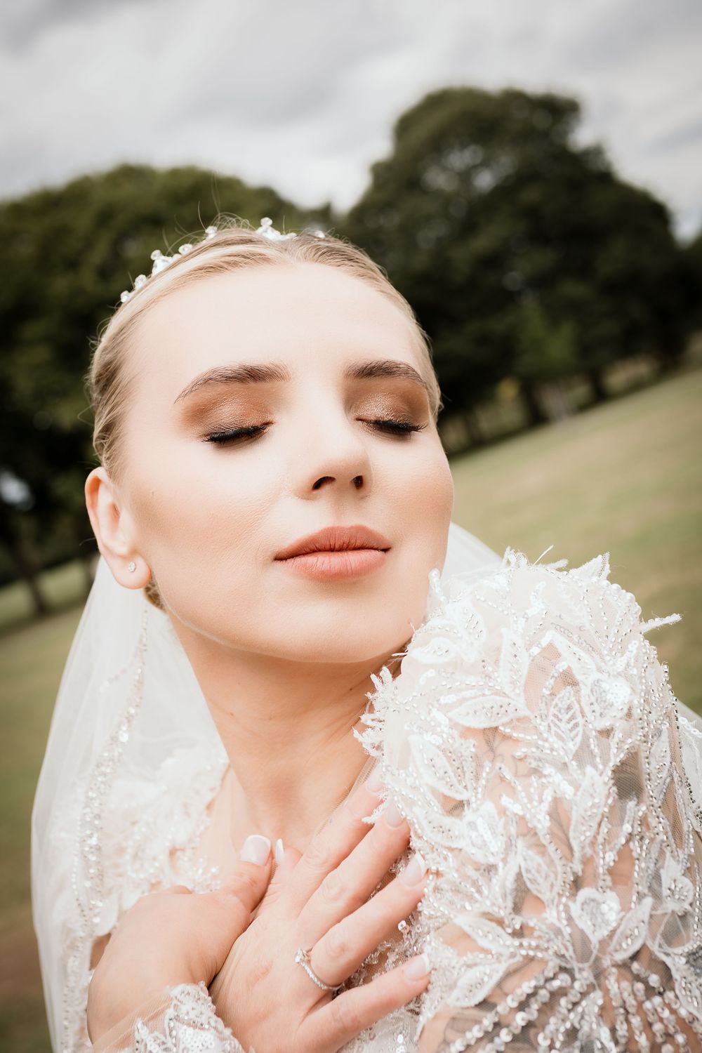 A bride wearing a delicate lace wedding dress and veil poses with closed eyes in an outdoor setting.