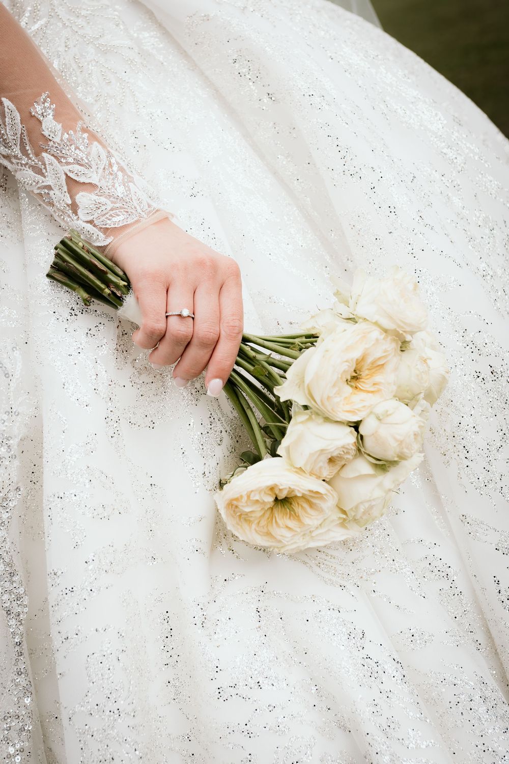 A hand in lace sleeve holding a bouquet of cream-colored roses against a white beaded wedding dress.