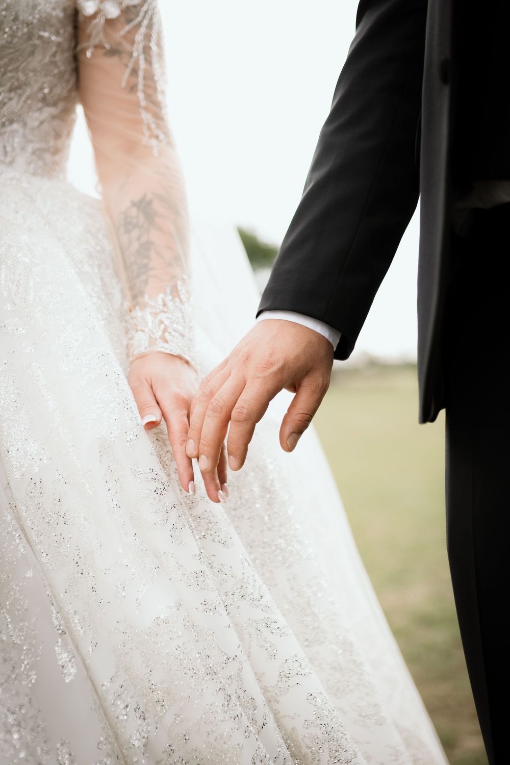Close-up of wedding couple holding hands, showing bride's white lace dress and groom's black suit sleeve.