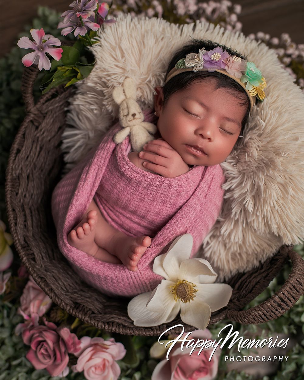 Sleeping newborn in pink wrap surrounded by flowers in wooden bowl.