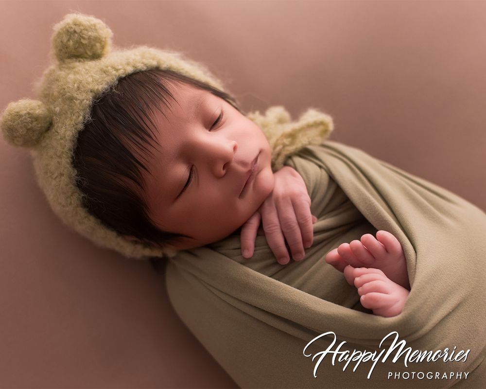 Sleeping newborn in beige wrap with teddy bear ears against warm backdrop.