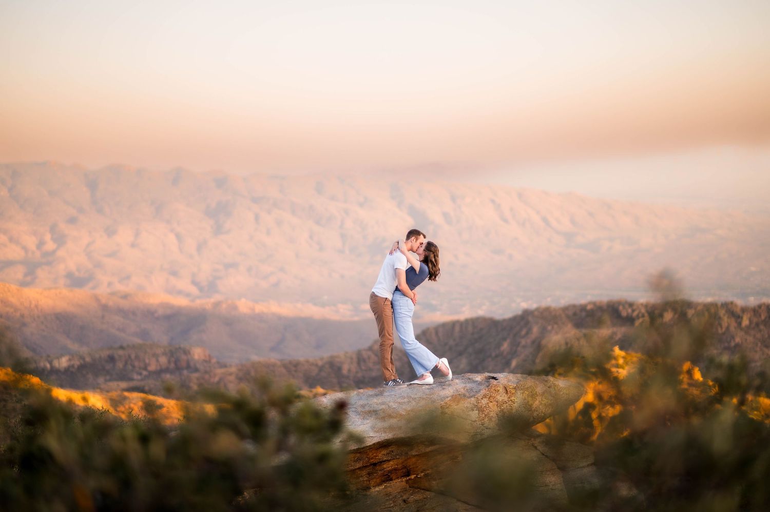 Romantic couple embracing on a rocky overlook with stunning mountain landscape at golden sunset.