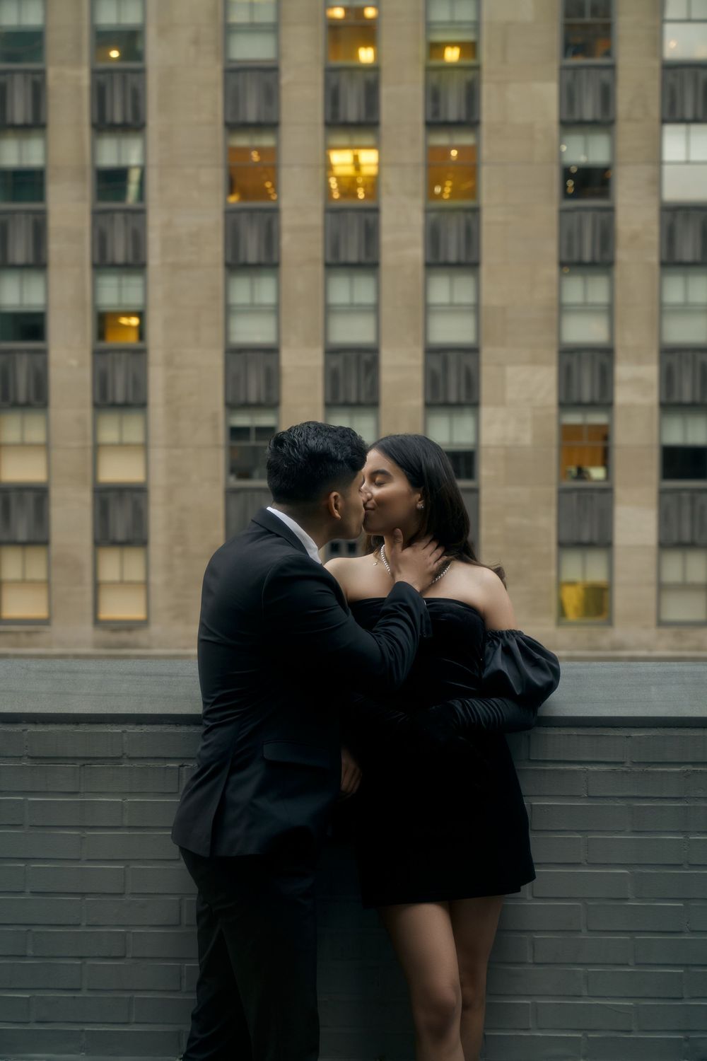 A romantic couple embraces and kisses against an urban backdrop of a tall building with illuminated windows at dusk by 3 West Club manhattan