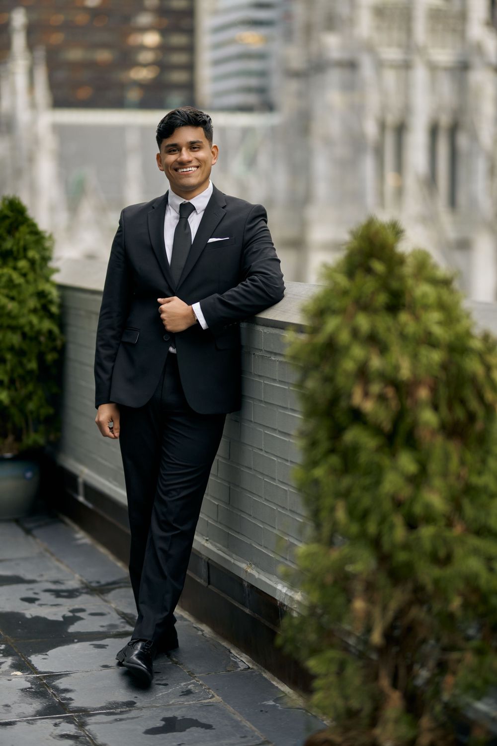 A well-dressed person in a black suit poses confidently against a gray wall next to green shrubbery by 3 West Club manhattan