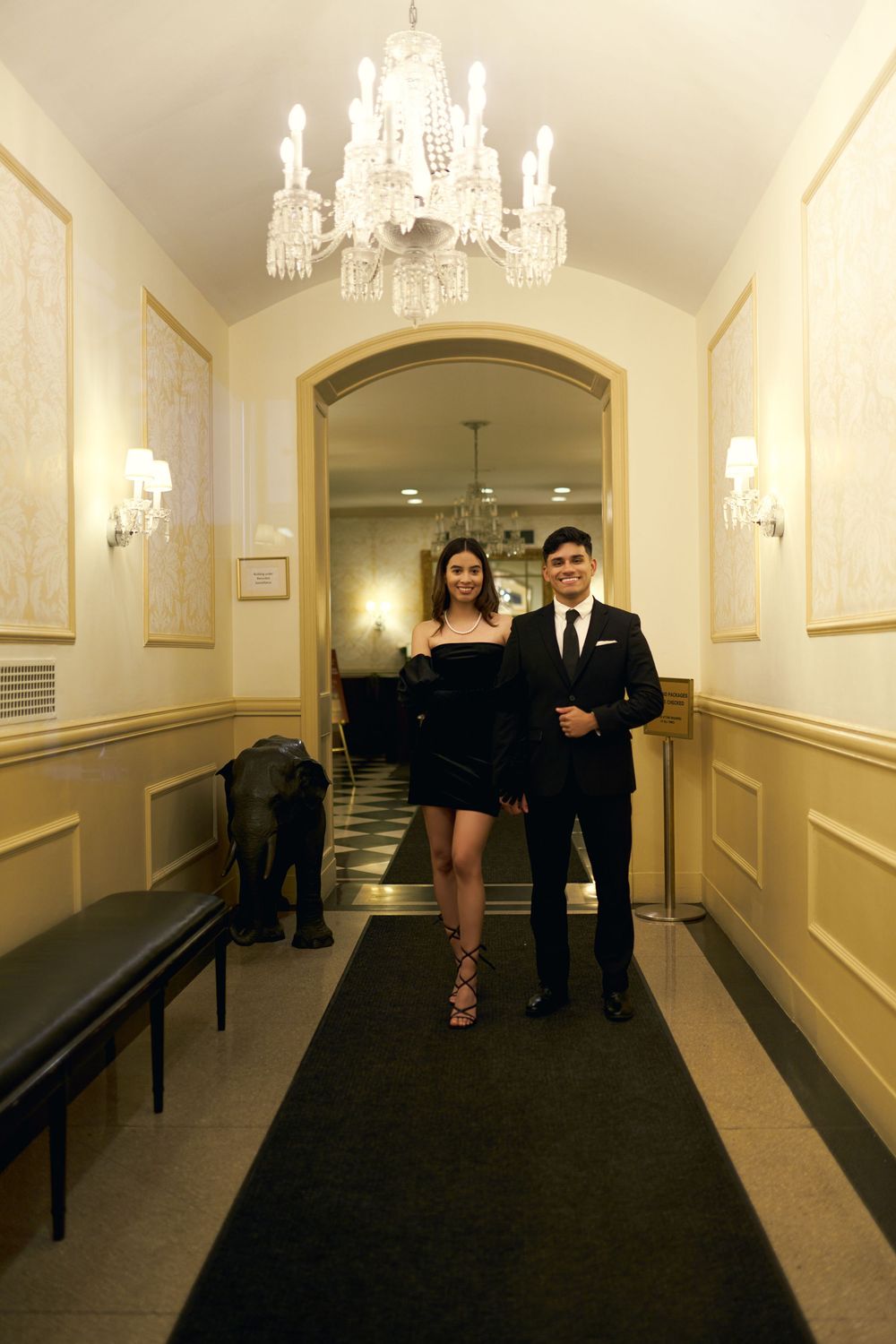 A formal couple walks down an elegant hallway with cream walls, wall sconces and a crystal chandelier overhead by 3 West Club manhattan