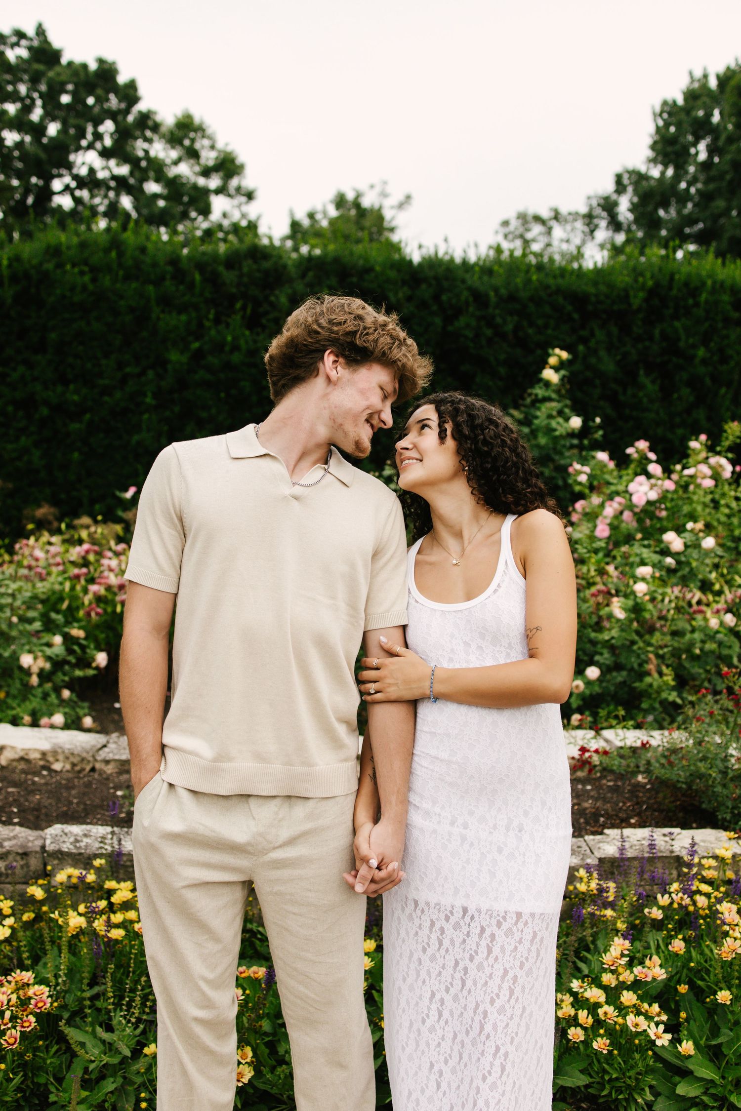 Couple in coordinated white and beige outfits posing in a garden with blooming flowers.